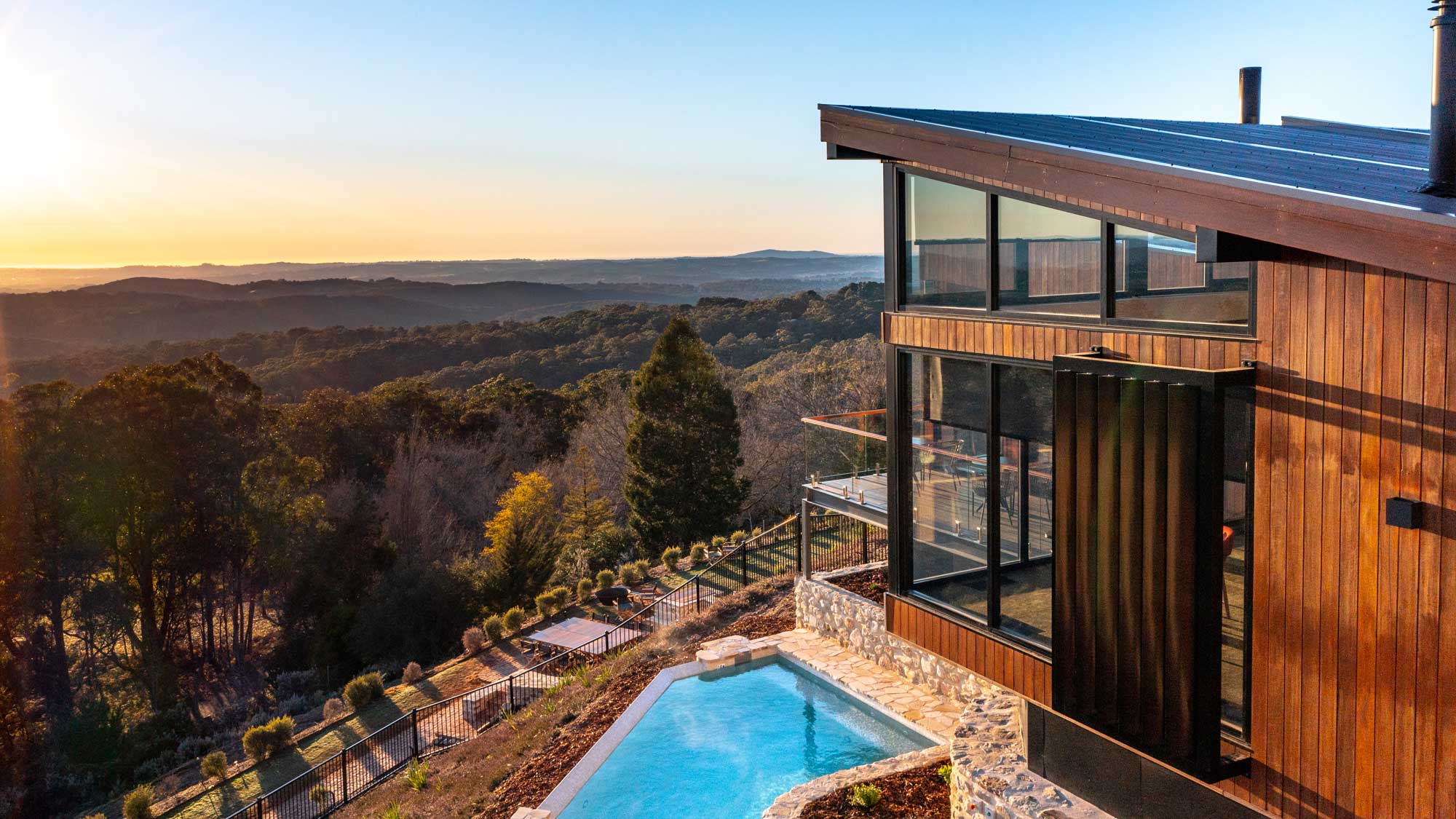 A modern wooden house with large glass windows overlooks a scenic landscape at sunset. A small pool is nestled beside the house, surrounded by greenery and hills in the distance.