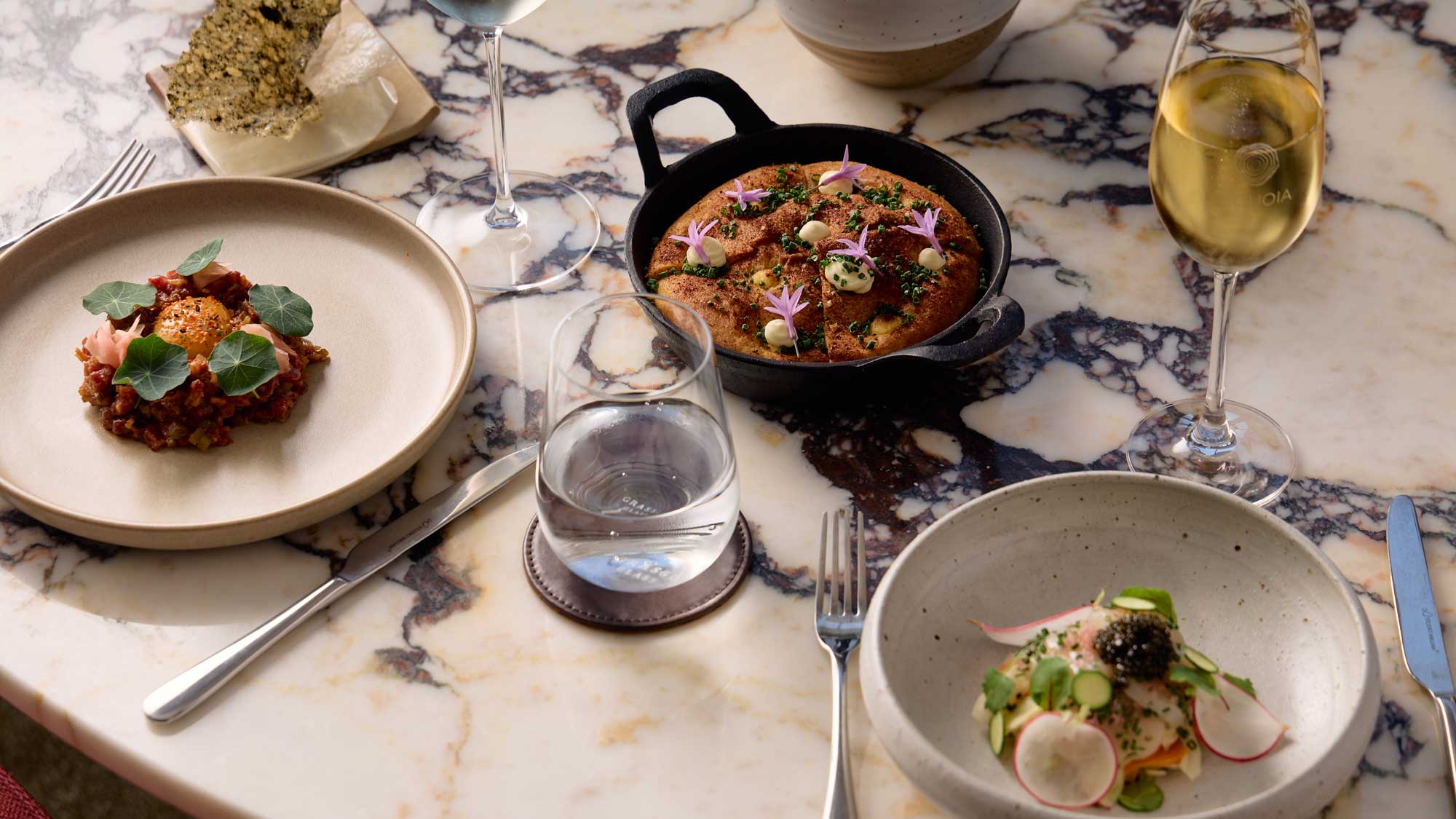 A beautifully arranged dining table featuring three gourmet dishes: a bowl with a vibrant seafood dish topped with microgreens and caviar, a skillet containing a savory rice dish adorned with edible flowers, and a plate with a rich meat dish garnished with fresh herbs. Two elegant glasses of sparkling wine and a water glass complete the setting on a marble table.