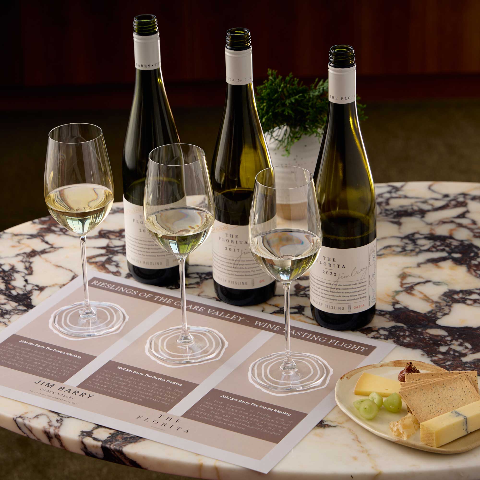 A marble table displays three elegant wine glasses filled with white wine, alongside four bottles of wine and a tasting menu titled "Rieslings of the Clare Valley." A plate with cheese and grapes is also present.