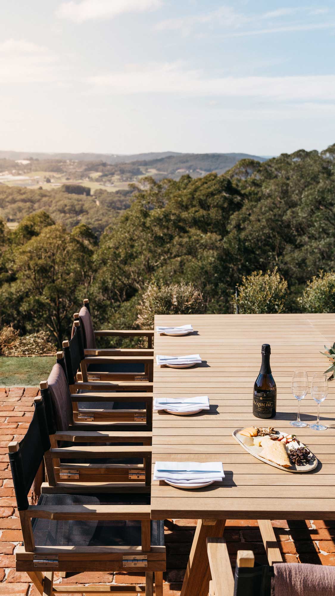 A wooden dining table set on a patio with several chairs, overlooking lush green hills and a scenic landscape. A bottle of wine and a platter of cheese and fruits are placed on the table, with glassware ready for use. The sun shines softly in the clear sky, highlighting the tranquil setting.