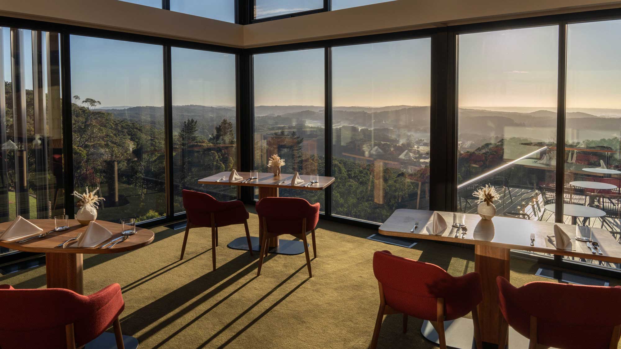 A modern restaurant interior featuring large glass windows with a scenic view of rolling hills and trees. The tables are elegantly set with white cloths and flowers, and plush red chairs surround the tables. Soft sunlight illuminates the space, creating a warm and inviting atmosphere.