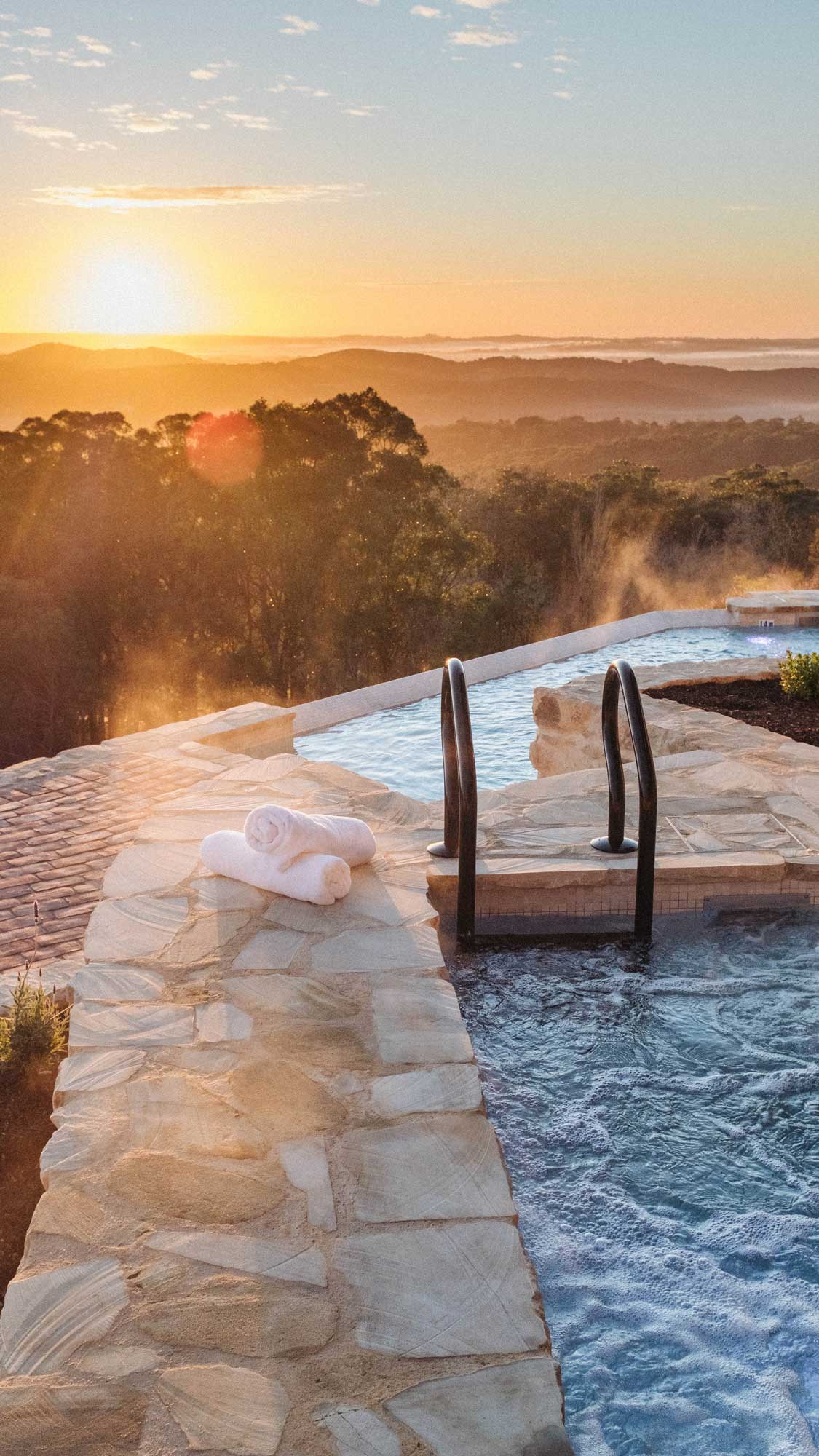 A serene outdoor hot tub overlooking a picturesque landscape at sunset. Rolling hills and a misty horizon are visible in the distance, with soft golden light bathing the scene. Two white towels are neatly placed on the stone pathway beside the tub.