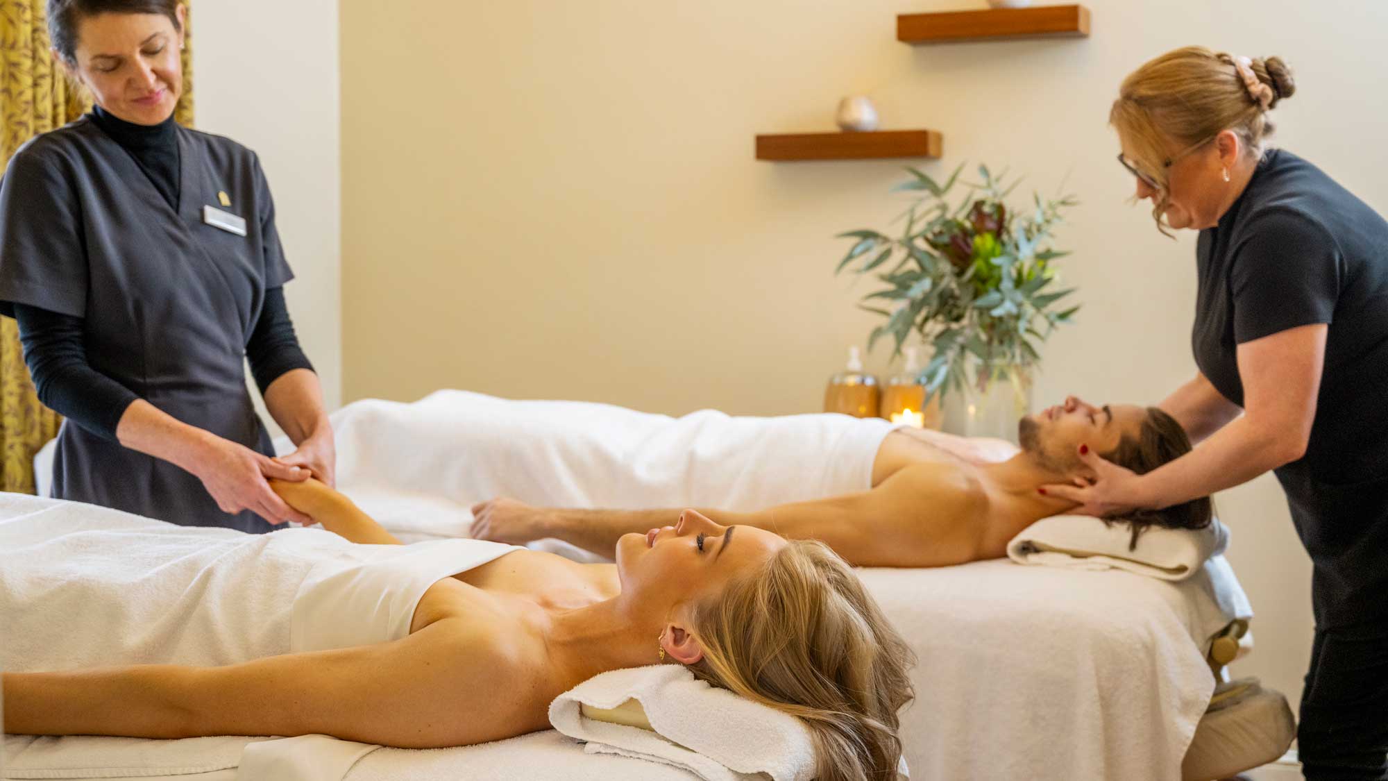 A serene spa setting with two people receiving massages on two treatment beds. A woman is focused on massaging the arm of a woman lying down, while another woman provides a neck massage to a man. Soft lighting and calming decor enhance the relaxing atmosphere.