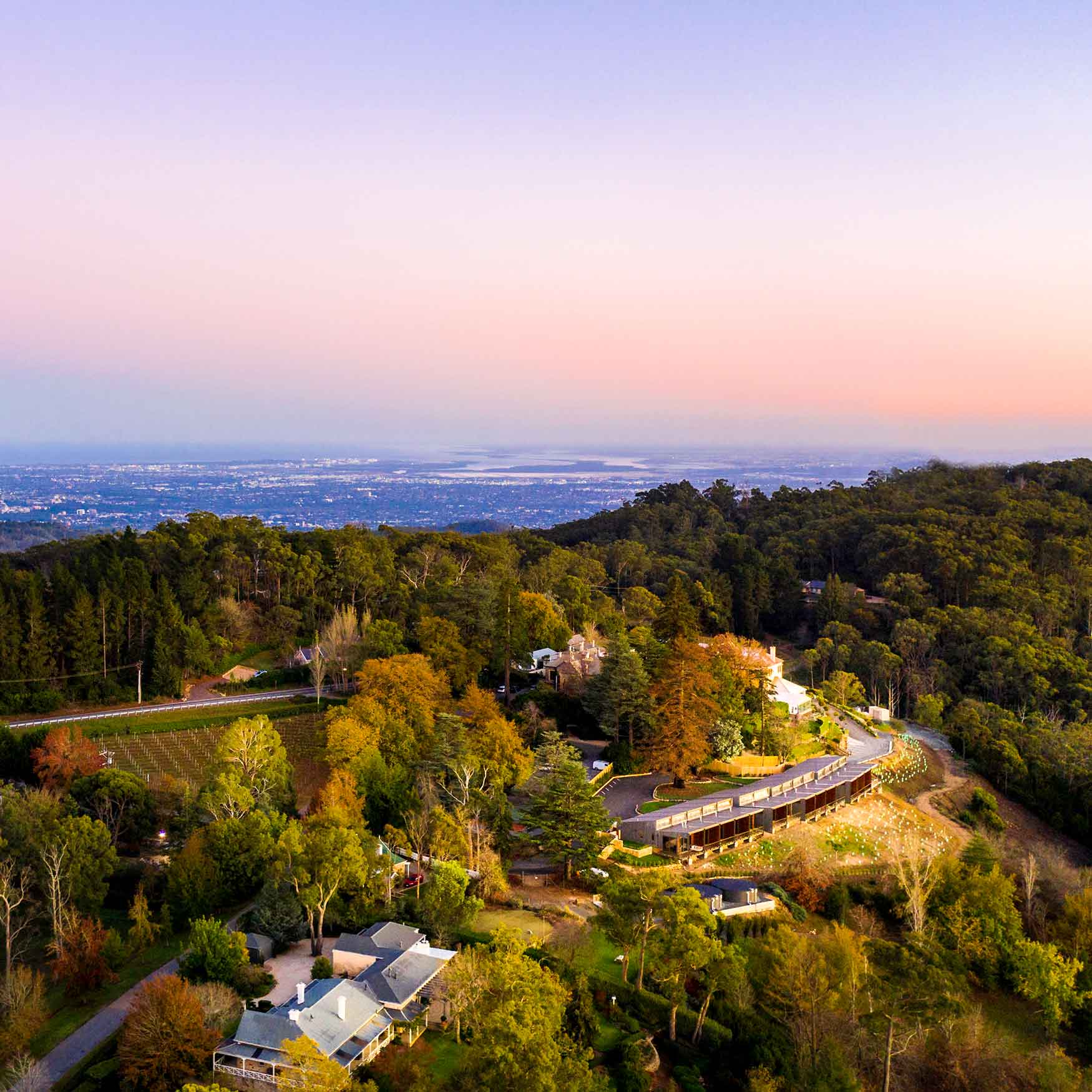 Aerial view of a hillside lodge surrounded by trees, with a vibrant sunset sky and a distant cityscape visible in the background.