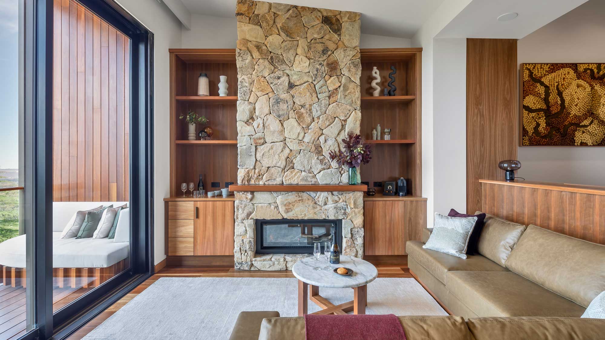 A modern living room features a large stone fireplace surrounded by wooden shelves. The space includes a cozy sofa with decorative pillows, a small round coffee table with a bowl of fruit, and large windows allowing natural light. A plush area rug complements the warm tones of the decor.