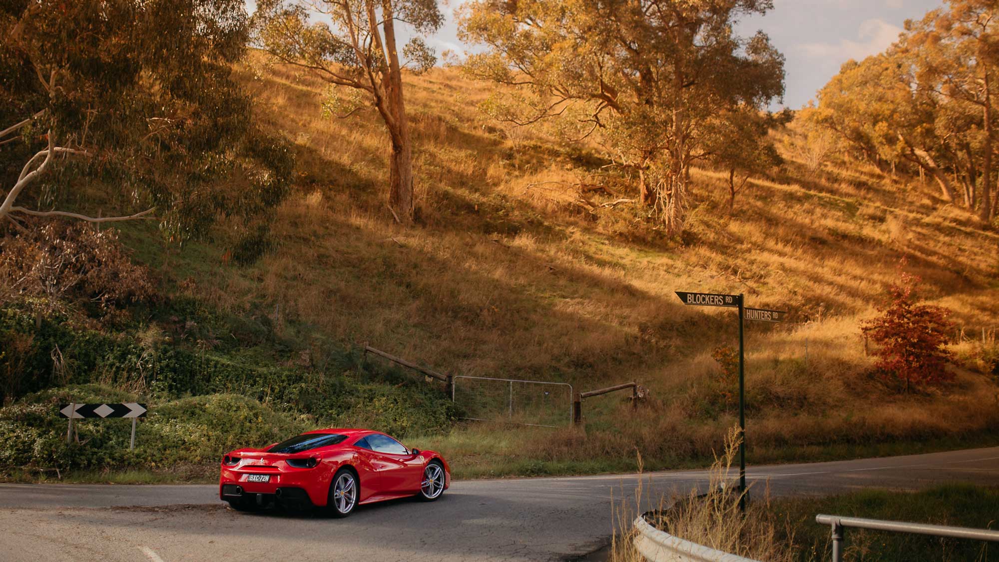 A striking red sports car navigates a winding road surrounded by lush greenery and rolling hills. Sunlight illuminates the scene, highlighting the curves of the road and the car's sleek design. A directional sign can be seen nearby, indicating various routes.