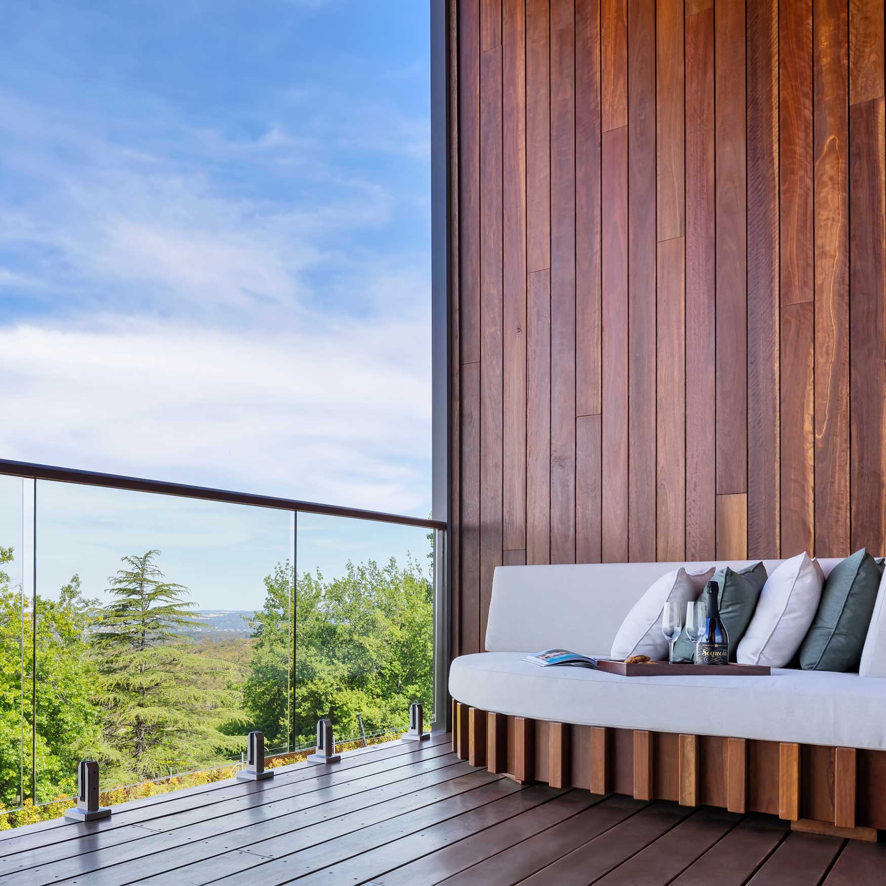 A modern balcony features a curved wooden seating area adorned with cushions. Glass railings provide a view of lush green trees and a bright blue sky.