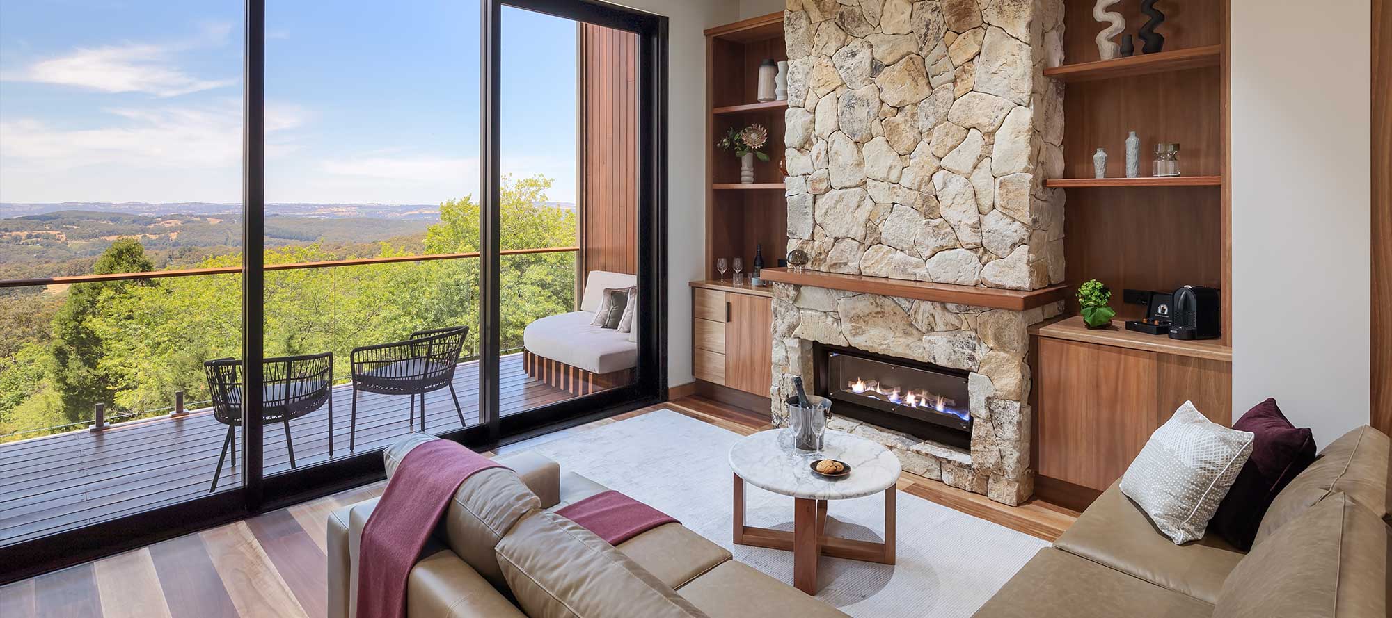 Modern living room featuring a stone fireplace, plush sofas with maroon pillows, and large glass doors opening to a scenic view of rolling hills. A round coffee table holds a decorative platter, alongside a sleek bookshelf with plants and accessories.
