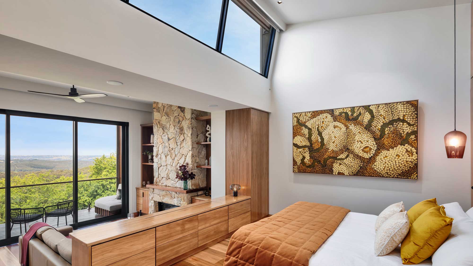 A modern bedroom featuring a large window with a scenic view. The room has a wooden dresser, a cozy bed with mustard-colored and white pillows, and a striking piece of wall art. A stone fireplace and a small seating area are also visible.