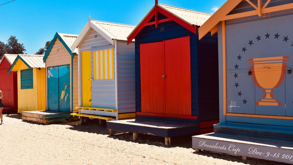 Colourful beach huts lined up on a sandy shore, featuring various bright designs and doors. The huts are adorned with artwork, and one is marked with the year "2019" and a trophy emblem.