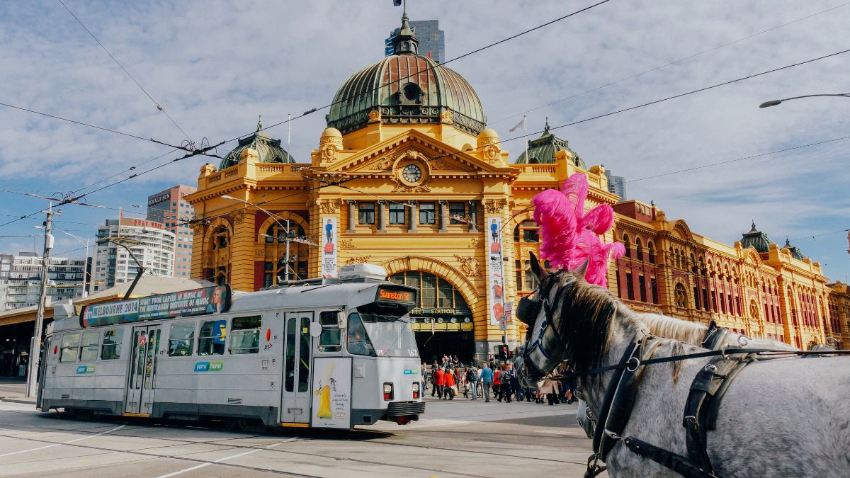 A horse wearing a pink feather plume stands in the foreground, while a tram passes by in front of the brightly colored Flinders Street Station in Melbourne. The vibrant architecture of the station is highlighted against a partly cloudy sky with urban buildings in the background.