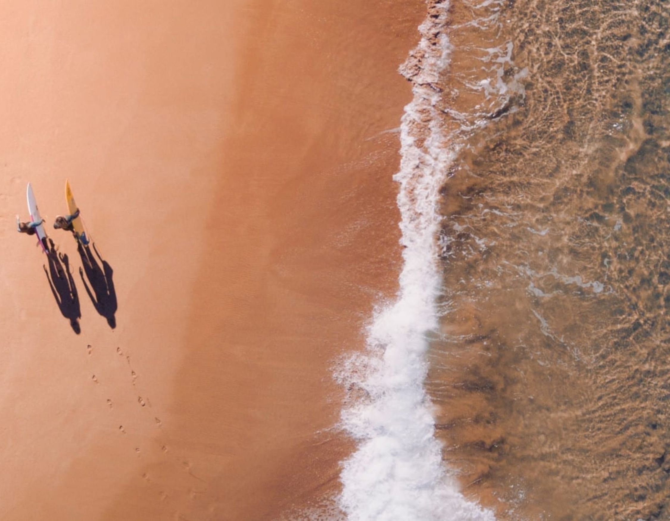 Two surfers walk along a sandy beach, carrying their surfboards. The gentle waves lap at the shore, creating a contrast between the water and the golden sand.