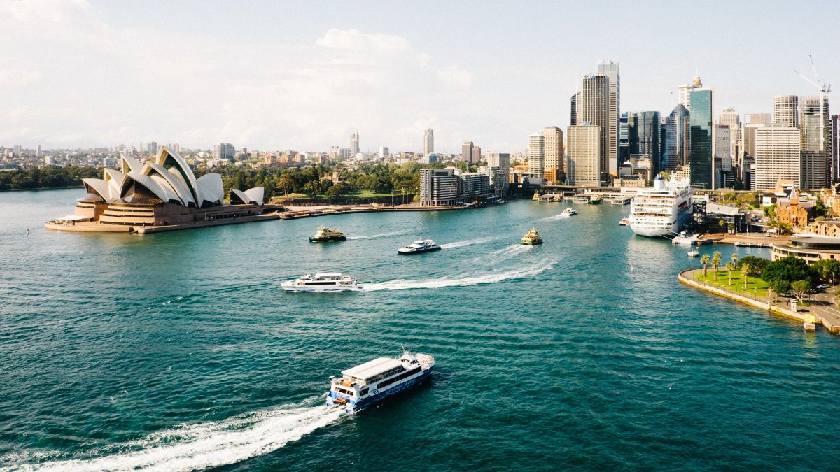 Sydney skyline with tall buildings and the iconic Sydney Opera House, surrounded by water and boats cruising in the harbor. Bright sunlight and a clear sky enhance the vibrant scene.