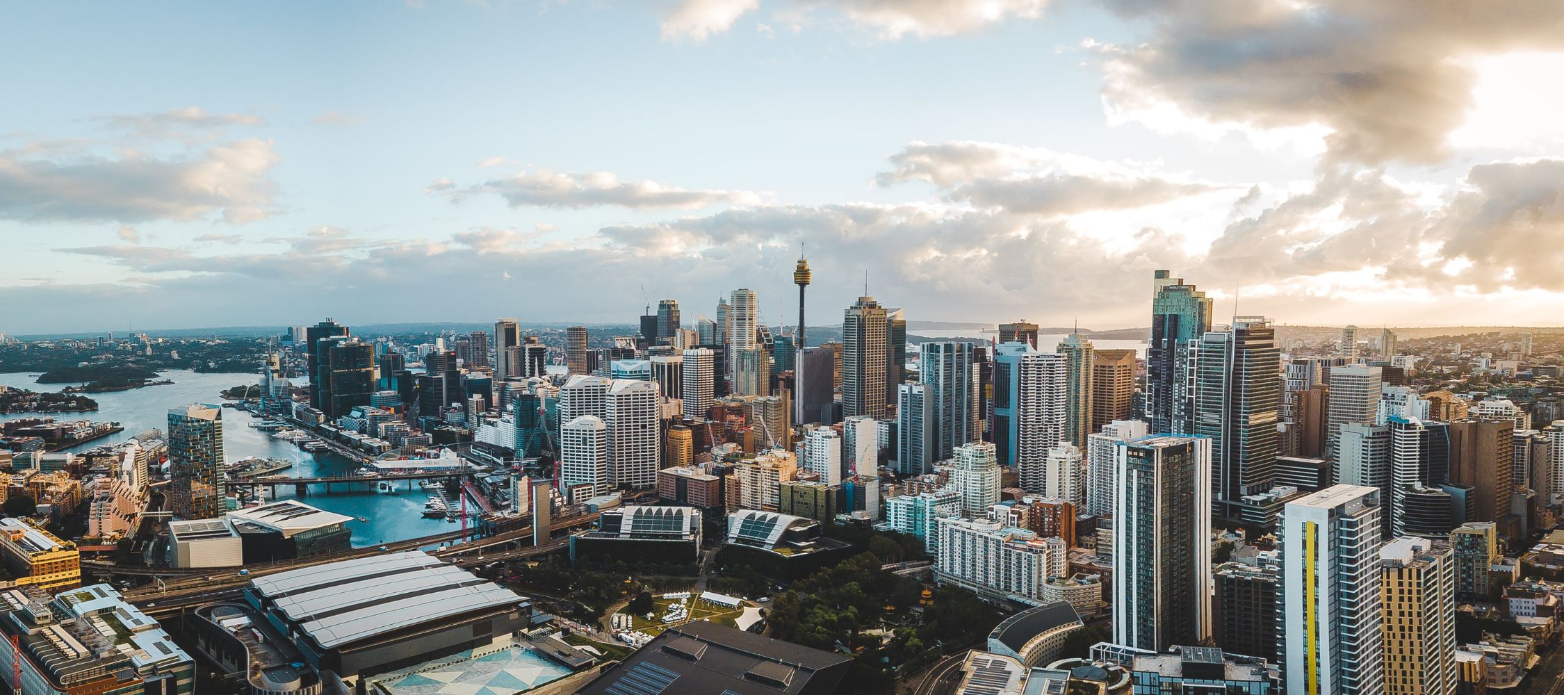 A panoramic view of a vibrant city skyline with modern skyscrapers, waterways, and a mix of clouds in the sky. The scene captures the contrast between urban architecture and natural elements, highlighted by a golden sunset.
