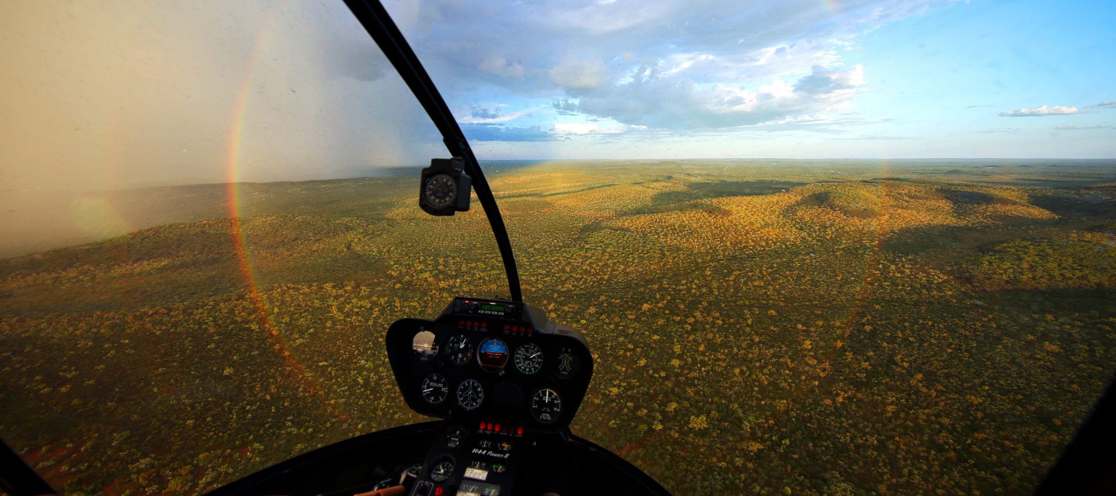 A panoramic view from inside a helicopter cockpit overlooking lush, rolling green hills under a partly cloudy sky. The landscape showcases a mix of vibrant greenery and hints of sunlight filtering through the clouds, creating a serene atmosphere.