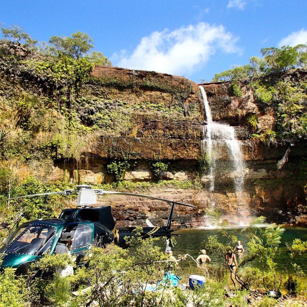A lush waterfall cascades down a rocky cliff into a serene pool surrounded by greenery. A helicopter is parked nearby, and a few people are enjoying the water. Blue sky with scattered clouds is visible above.