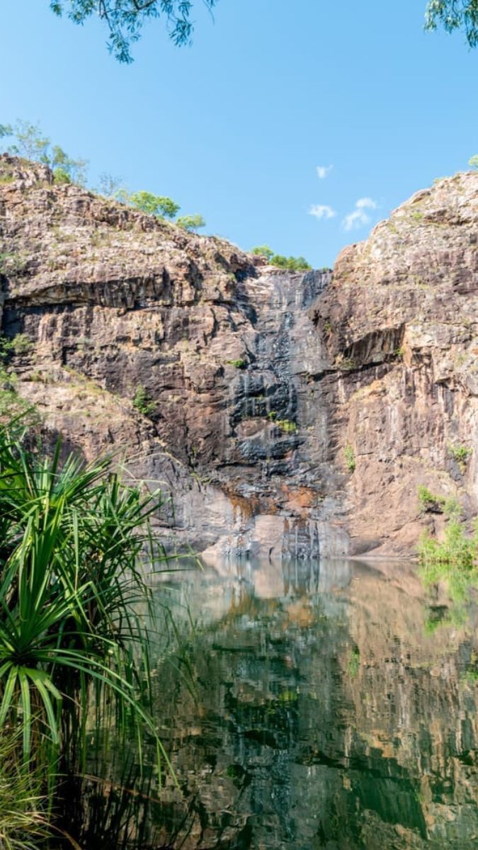 A serene waterfall cascading down rocky cliffs into a calm pool, surrounded by lush green vegetation under a clear blue sky.