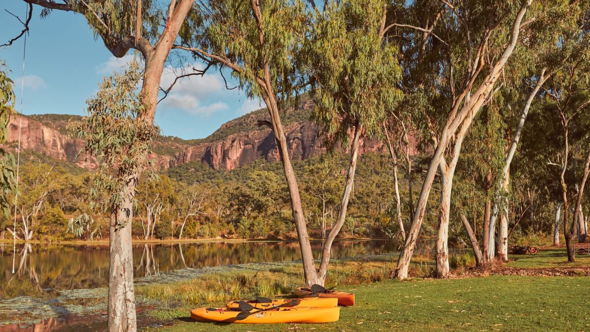 Two orange kayaks are resting on the grass near a calm lake surrounded by tall eucalyptus trees. In the background, rocky cliffs rise above lush vegetation under a clear blue sky.