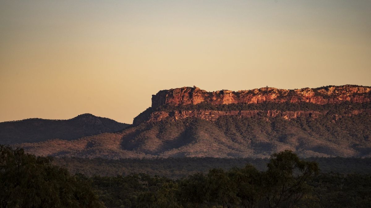 Sunset over a rugged, layered cliff with a flat top, surrounded by rolling hills and dense vegetation. The warm hues of the sky cast a soft glow on the landscape.