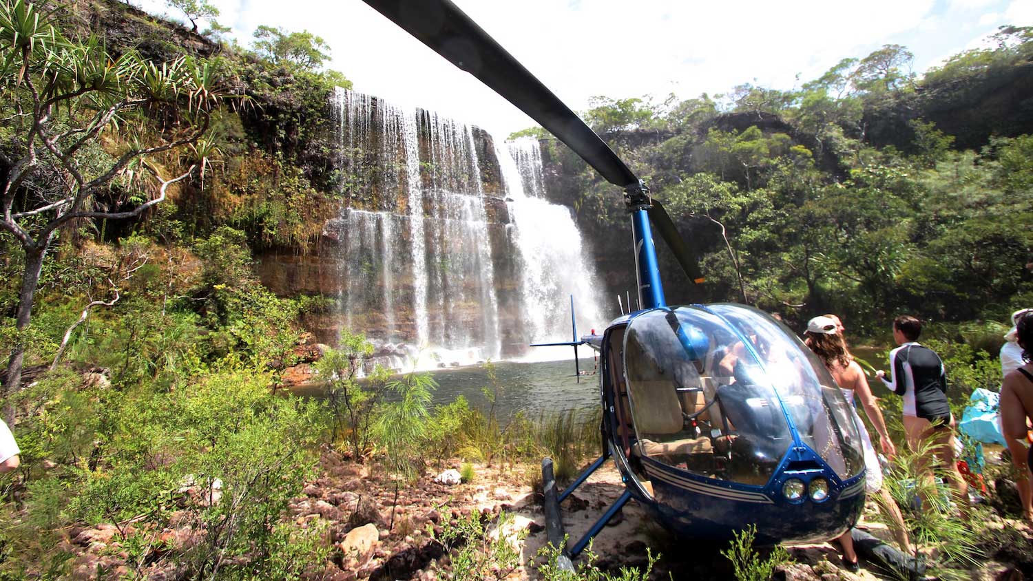 A blue helicopter parked near a lush waterfall surrounded by greenery, with a group of people admiring the scenery.