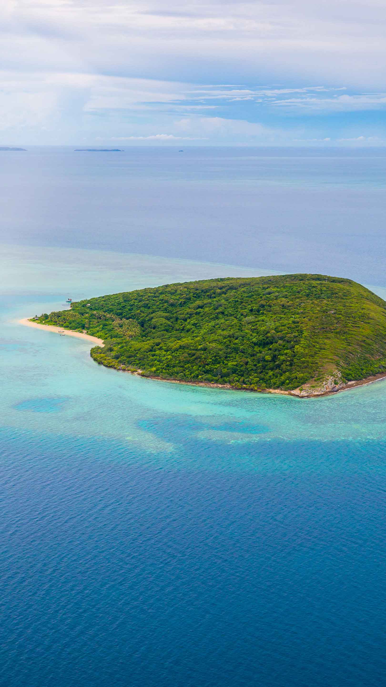 Aerial view of a lush, green island surrounded by turquoise waters and a sandy beach. The sky is partly cloudy, enhancing the serene landscape.