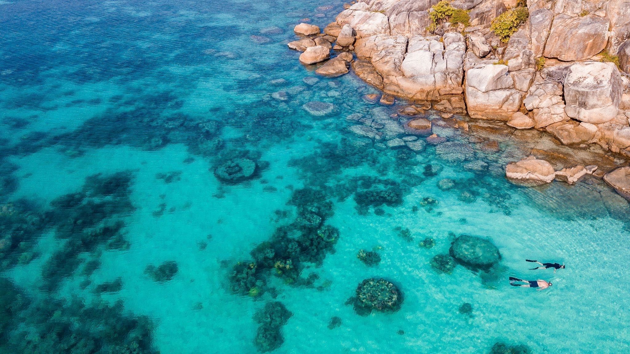 Aerial view of two snorkelers exploring clear turquoise waters near a rocky coastline, with visible coral formations and underwater textures.