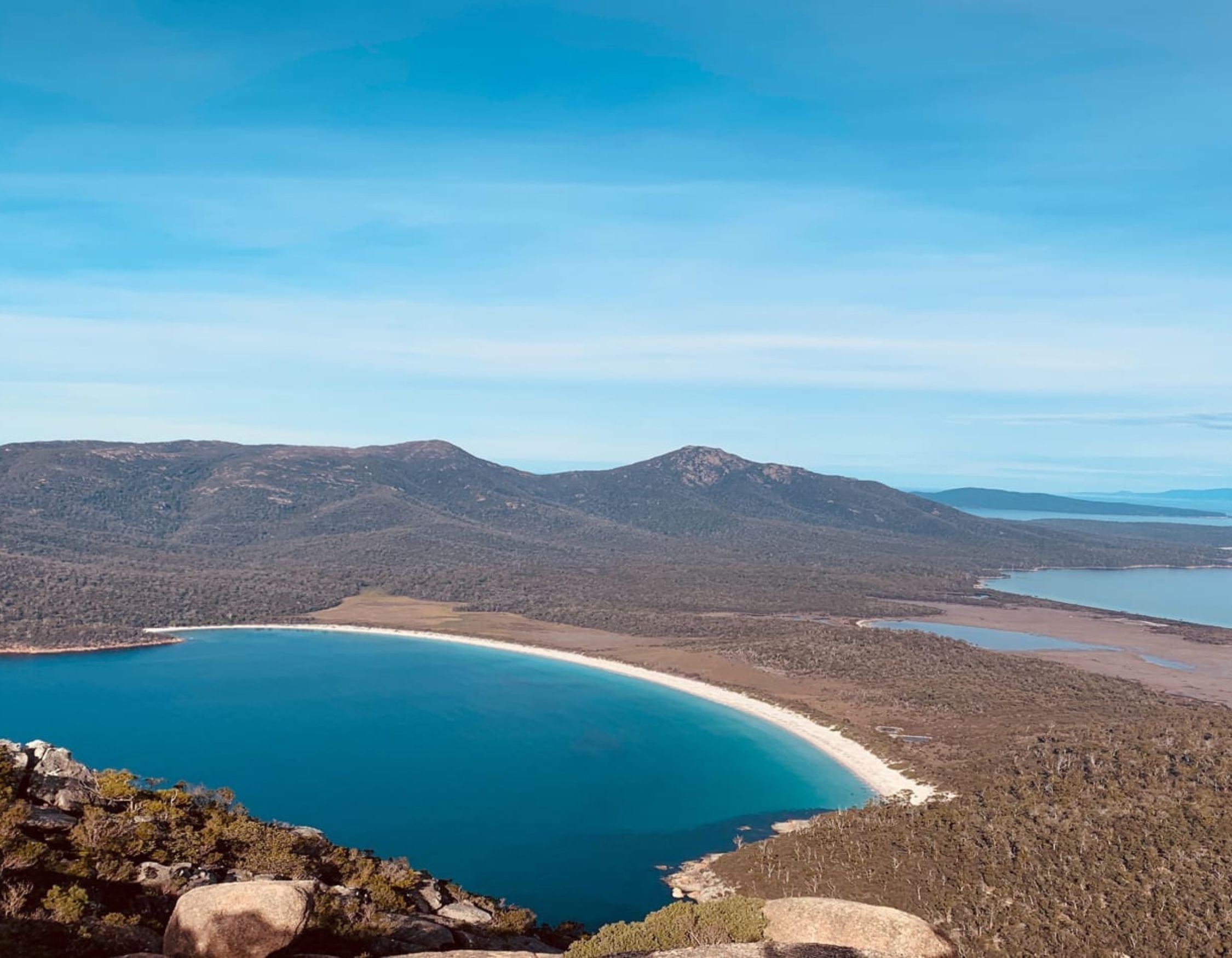 A panoramic view of a curved coastline bordered by calm blue waters, with lush green landscapes and distant mountains under a clear blue sky.