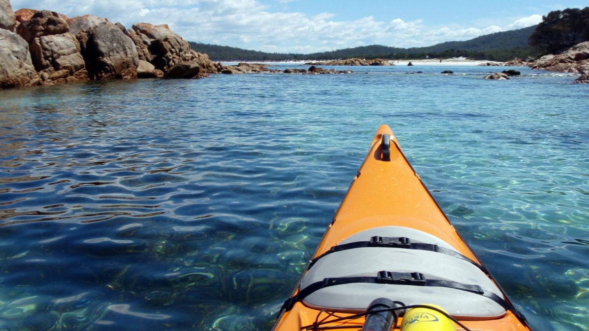 A bright orange kayak floats on clear, blue water, with rocky formations visible in the background and lush green hills in the distance. The sunlight reflects off the water's surface, creating a serene atmosphere.