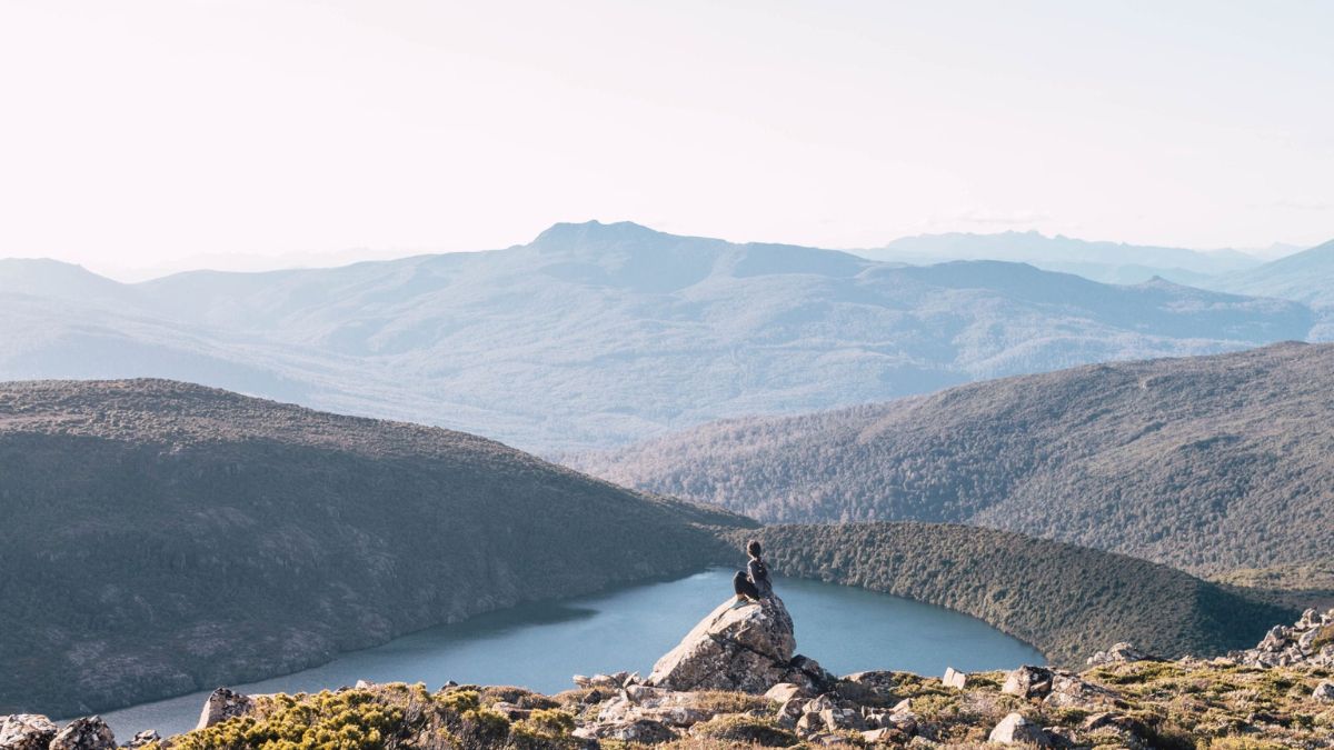 A person sitting on a rock overlooking a serene lake surrounded by lush mountains and valleys in the background. The scene is bathed in soft sunlight, creating a peaceful atmosphere.