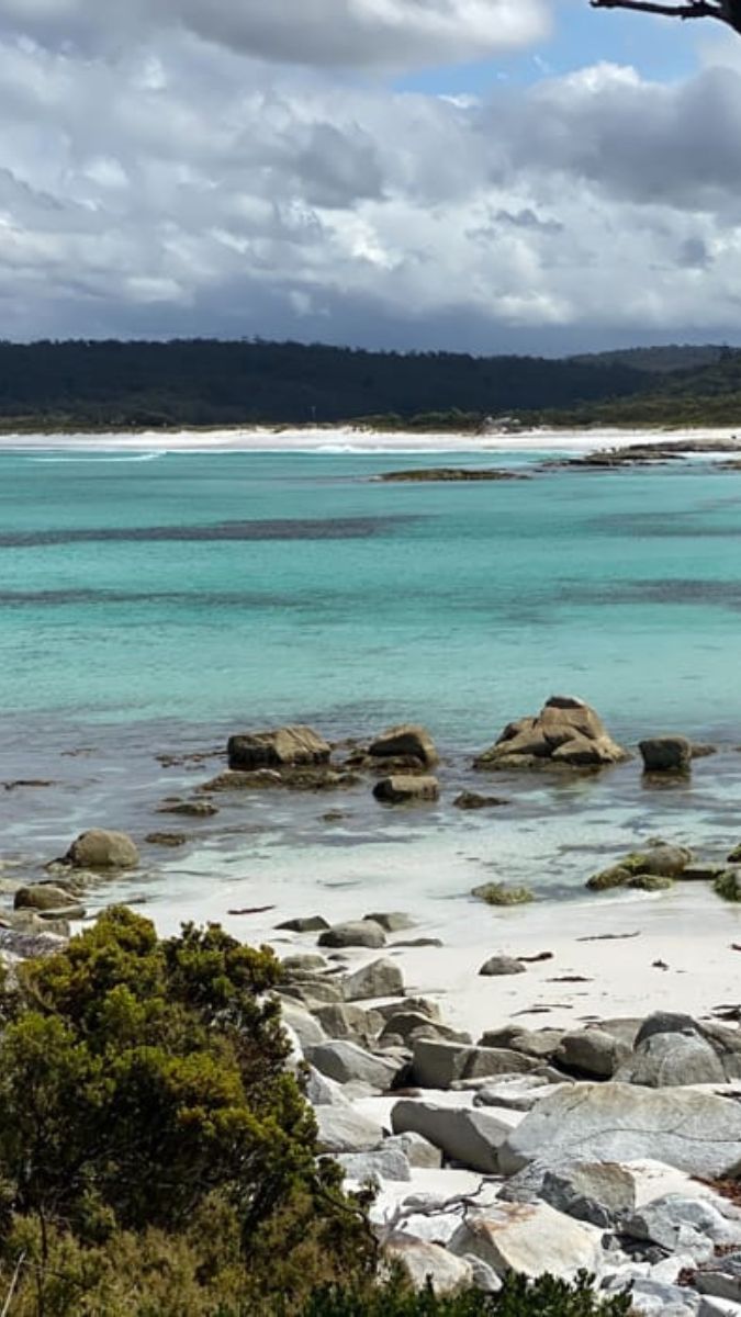 A serene coastal scene featuring turquoise waters gently lapping against rocky shores. Lush greenery borders the shoreline, with distant hills under a cloudy sky.