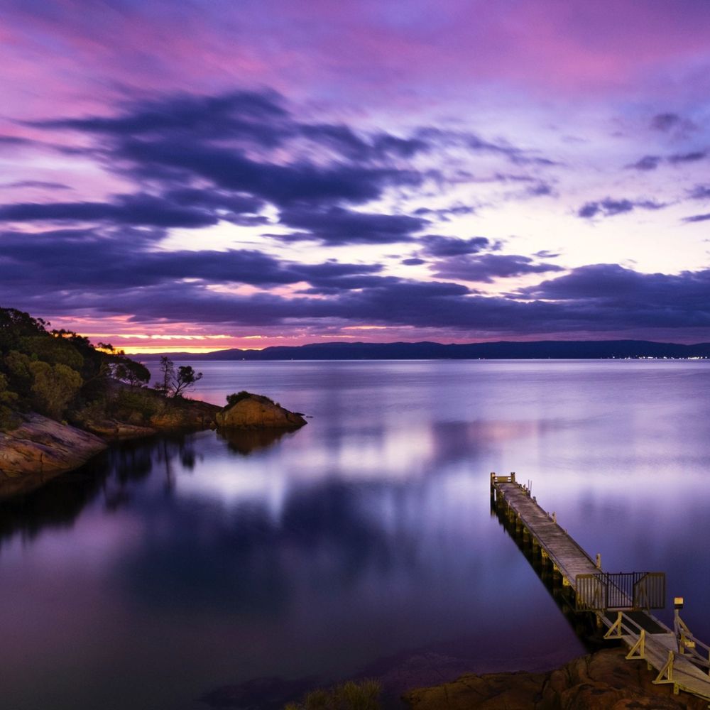 A serene waterfront scene at dusk featuring a calm body of water reflecting vibrant pink and purple hues in the sky. A wooden dock extends into the water, surrounded by lush greenery and distant hills.