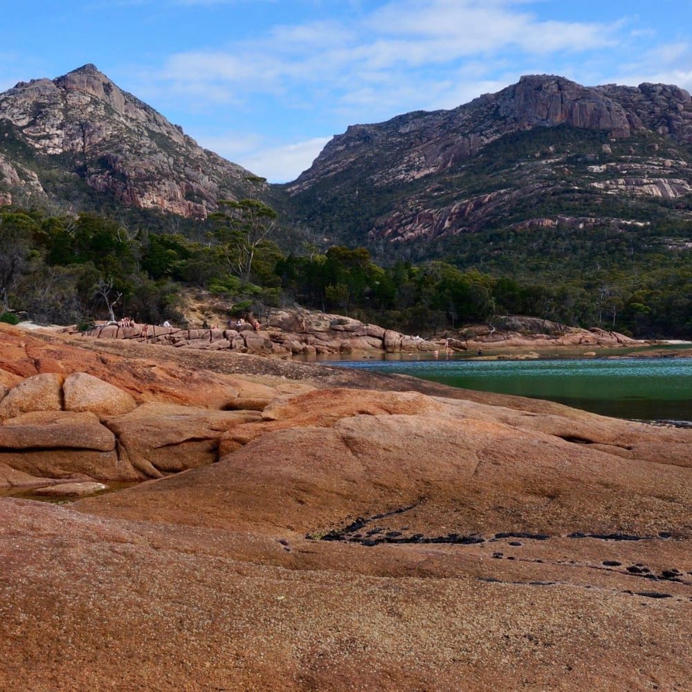 Rocky shoreline with large boulders in the foreground, leading to a tranquil bay surrounded by lush green trees and steep mountains in the background under a partly cloudy sky.
