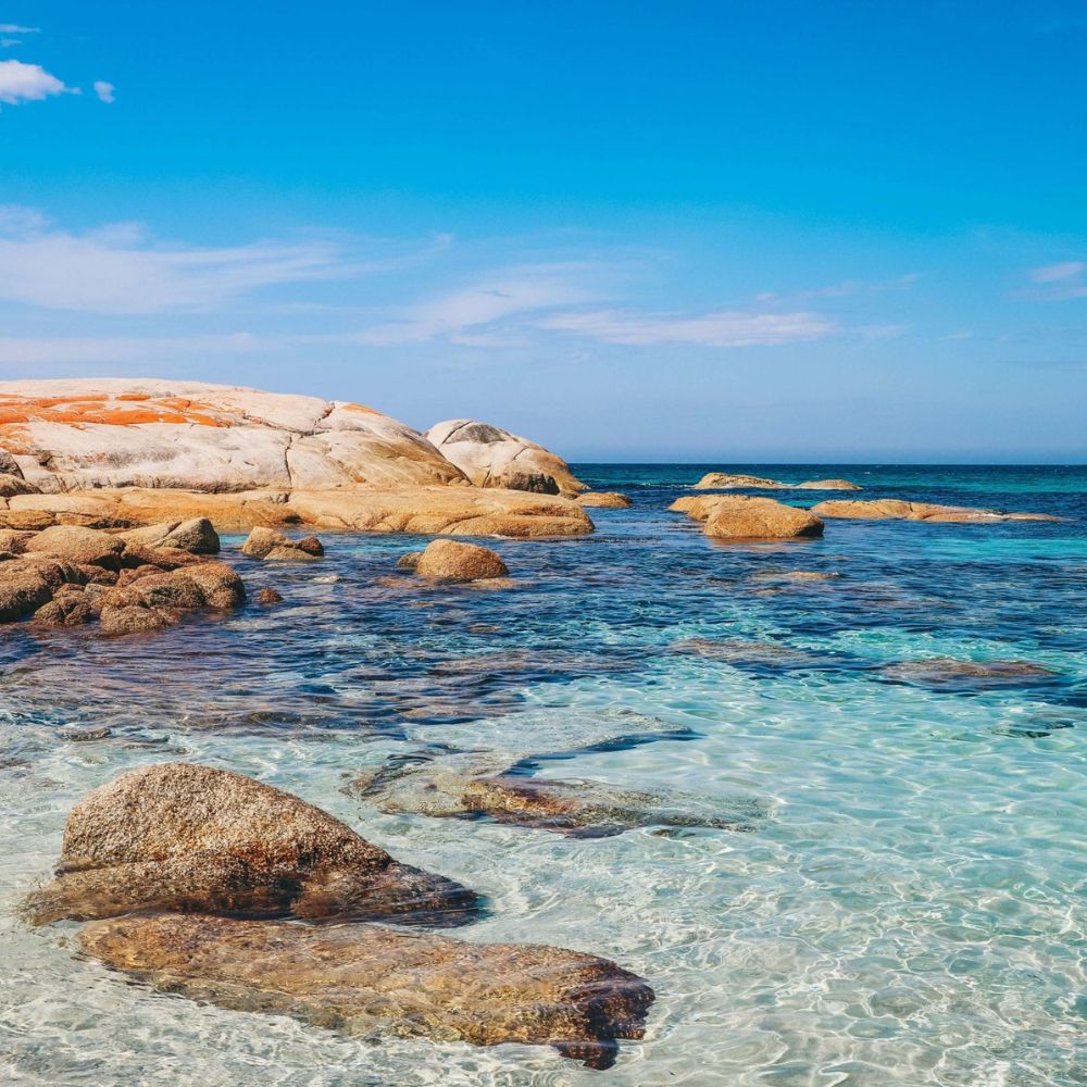 Clear turquoise waters gently lap against smooth, rocky shores under a bright blue sky. Sunlit boulders are scattered throughout the shallow water, creating a picturesque coastal scene.