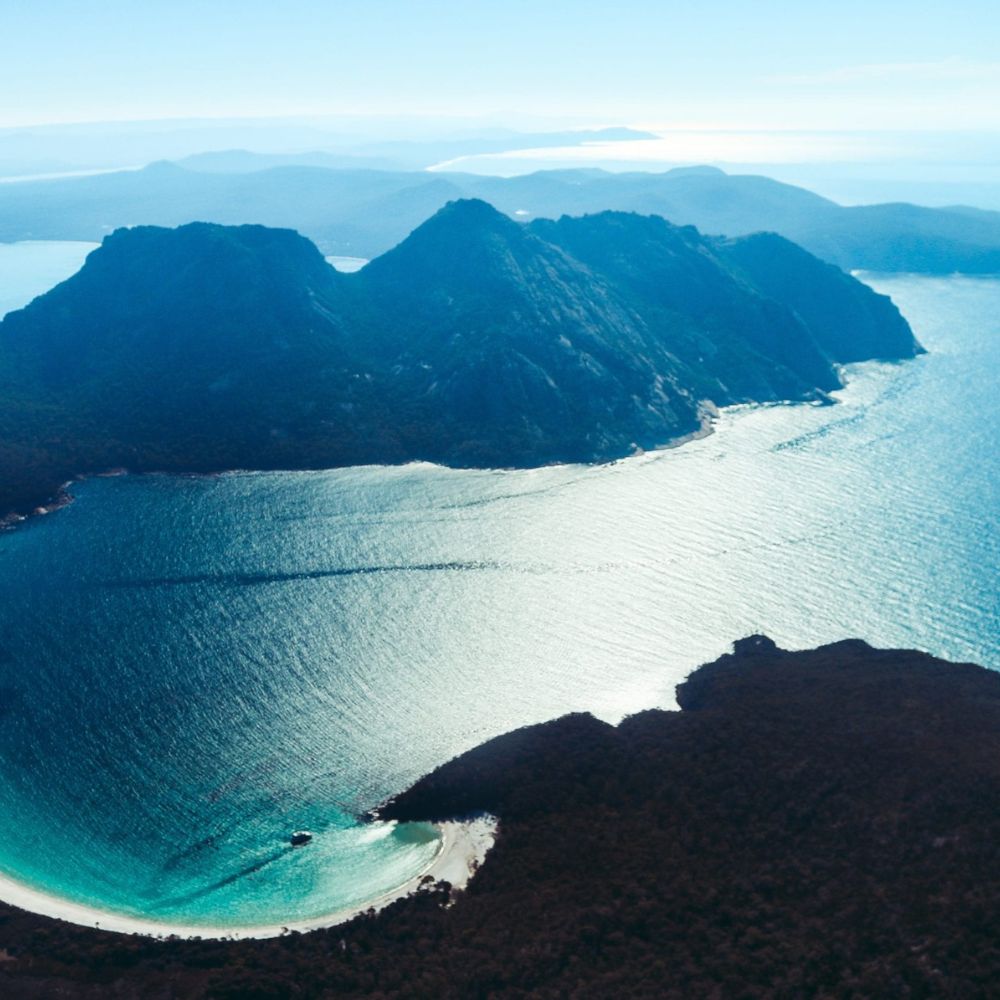 Aerial view of a rugged coastline with mountains in the background and sparkling blue waters. Gentle waves lapping at a sandy beach surrounded by lush greenery.
