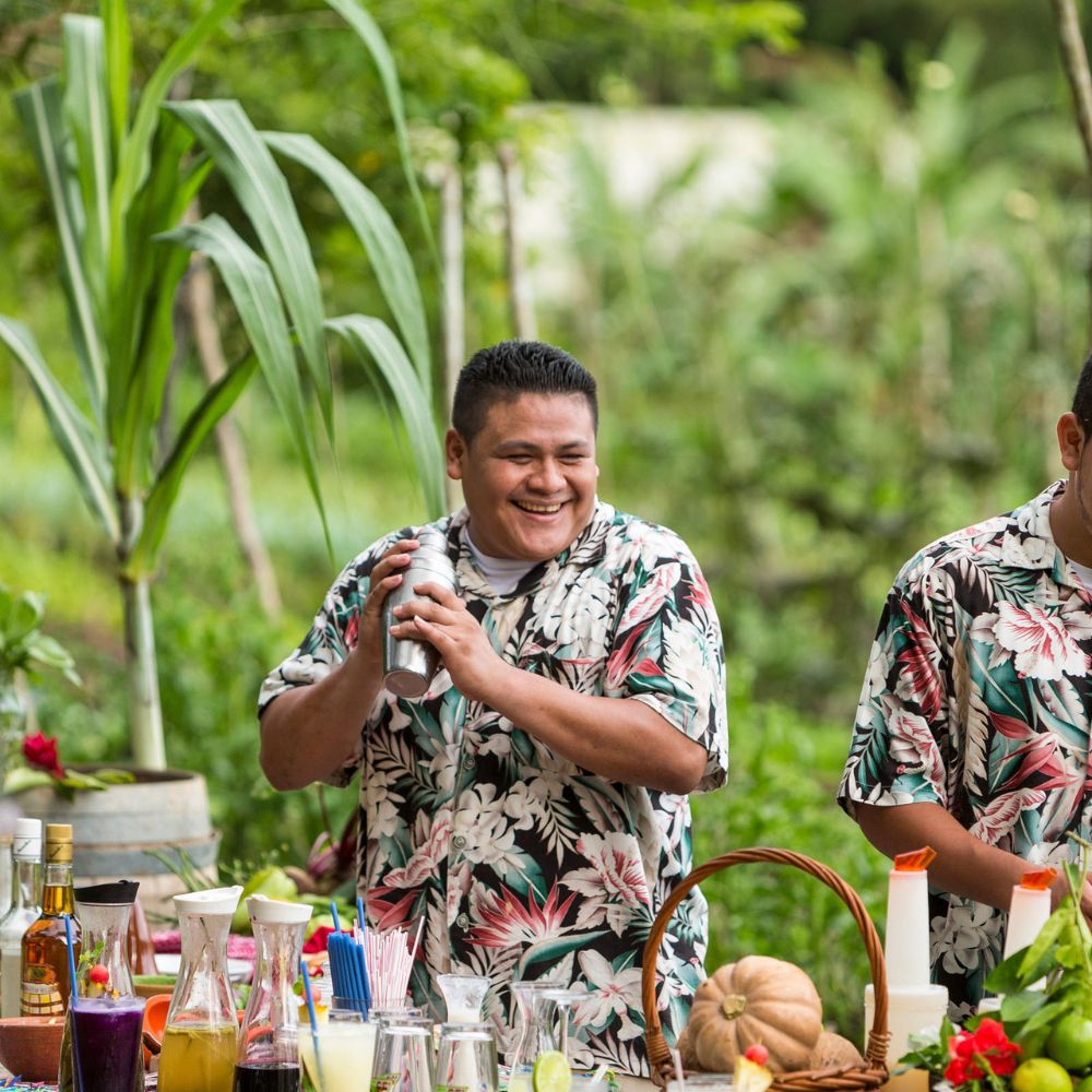 A cheerful person in a floral shirt shakes a cocktail shaker, surrounded by colorful drink ingredients and tropical greenery.