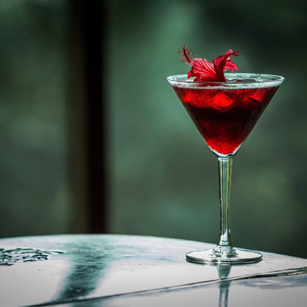 A vibrant red cocktail garnished with a hibiscus flower, placed on a reflective surface.