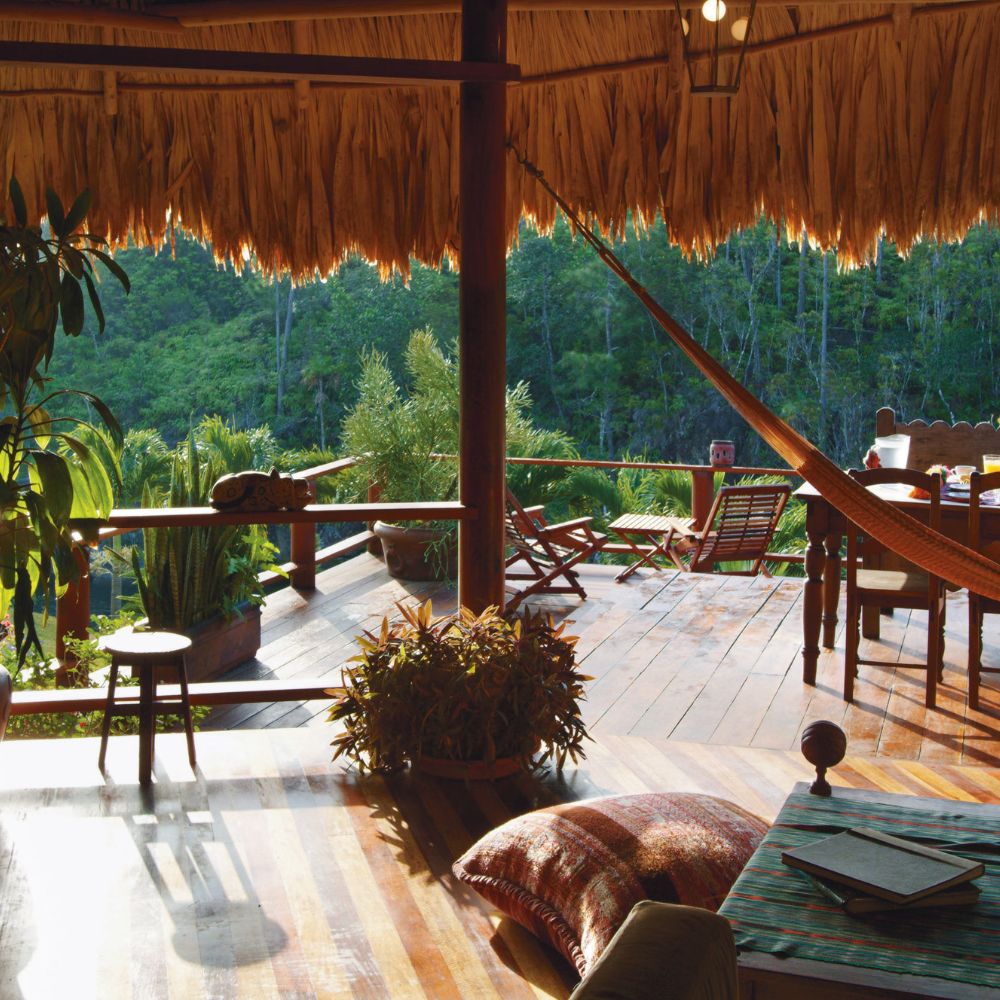 A cozy wooden deck with a thatched roof overlooking lush greenery, featuring a hammock, comfortable seating, and a small table. Sunlight filters through, creating a warm atmosphere.