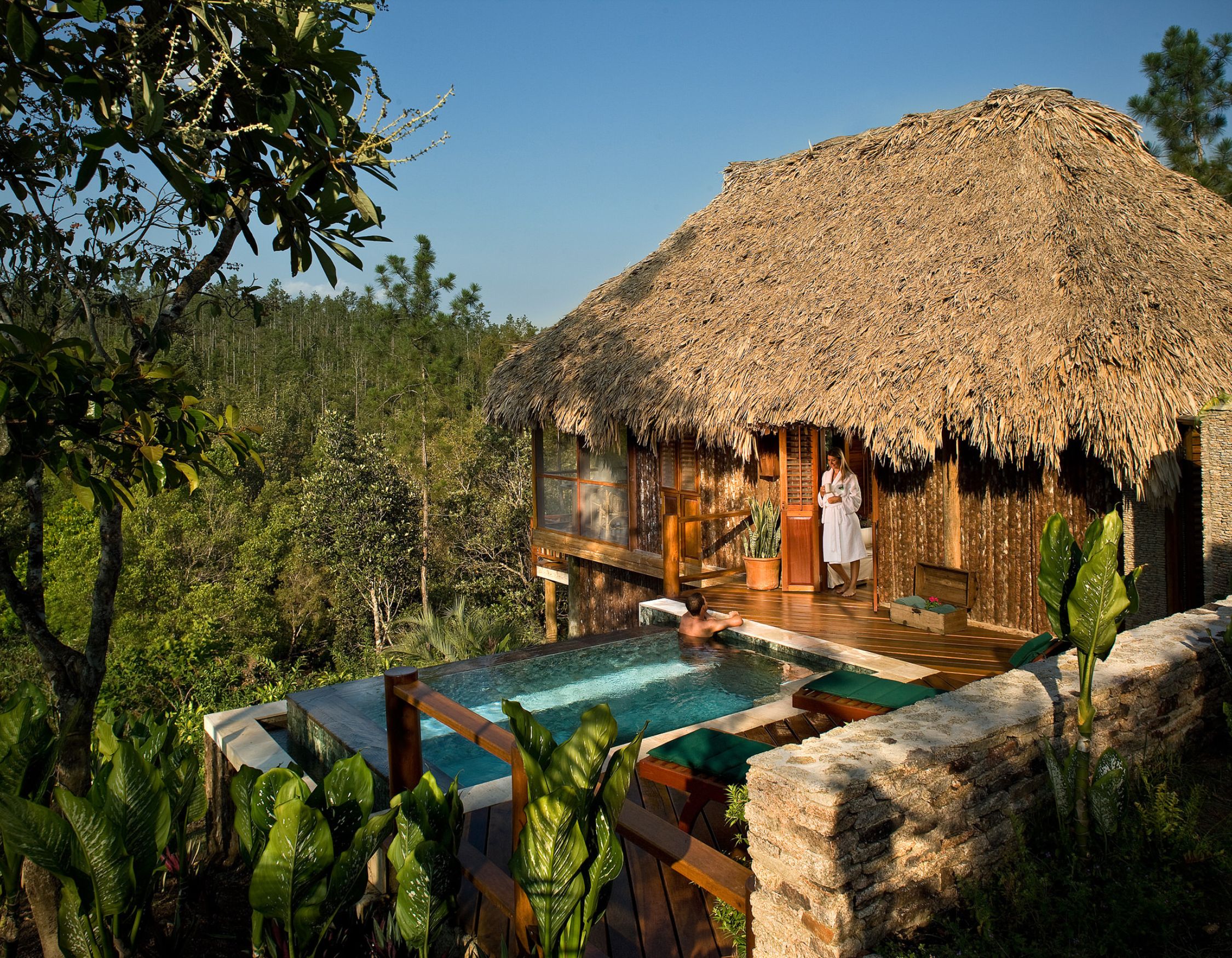 Luxury thatched cabin surrounded by lush greenery, featuring a private pool and wooden deck. A person stands on the balcony, while another relaxes in the pool.