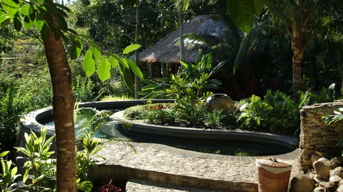 A serene garden featuring a winding pathway beside lush greenery, tropical plants, and a small thatched-roof hut in the background. A decorative pot rests among the foliage, enhancing the tranquil atmosphere.