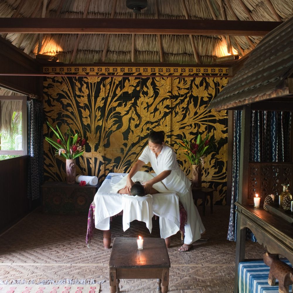 A serene spa room with a therapist giving a massage to a client on a massage table. The walls are decorated with intricate, dark floral designs, and tropical plants add a touch of nature. Soft lighting creates a calming atmosphere.