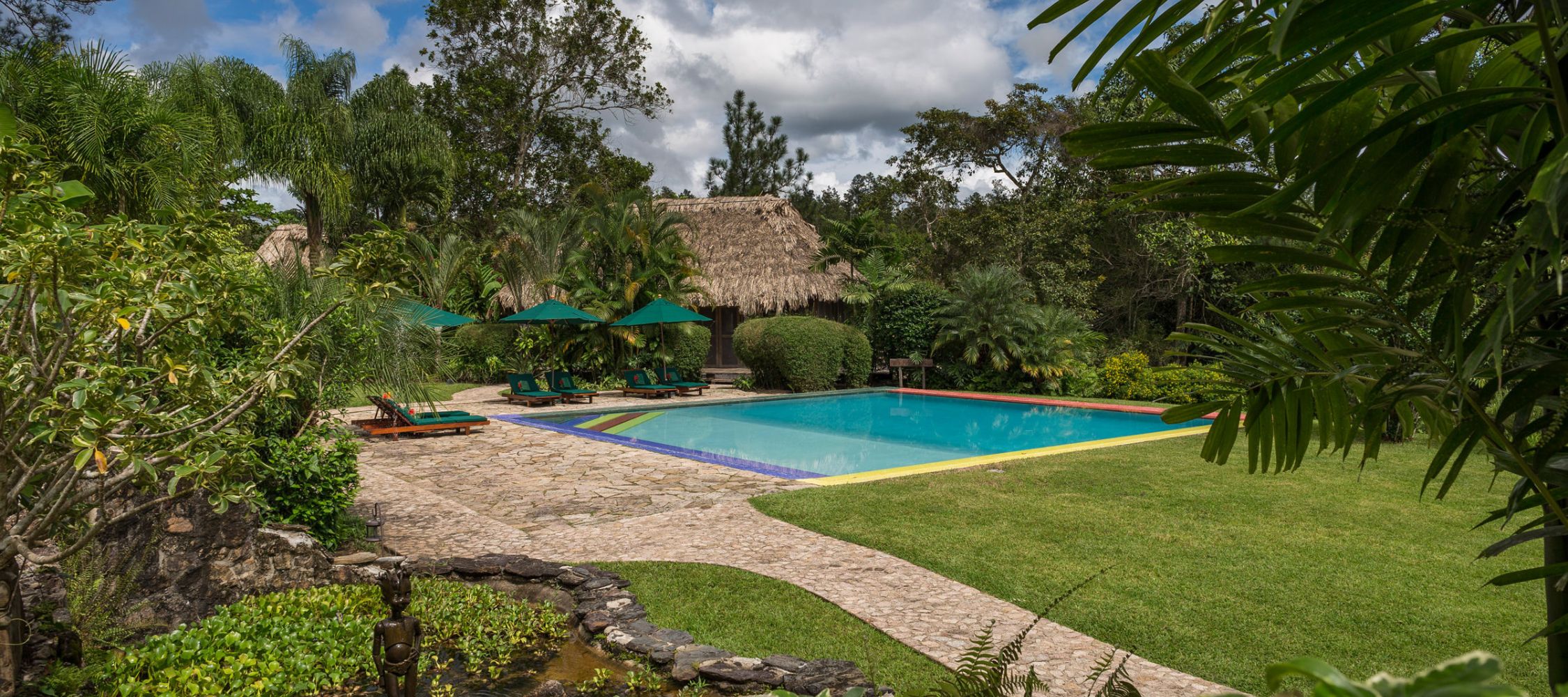 A tranquil outdoor scene featuring a swimming pool surrounded by lush greenery and tropical plants. Palapa-style huts are visible in the background, with lounge chairs and umbrellas arranged by the poolside. The sky is partly cloudy, creating a serene atmosphere.