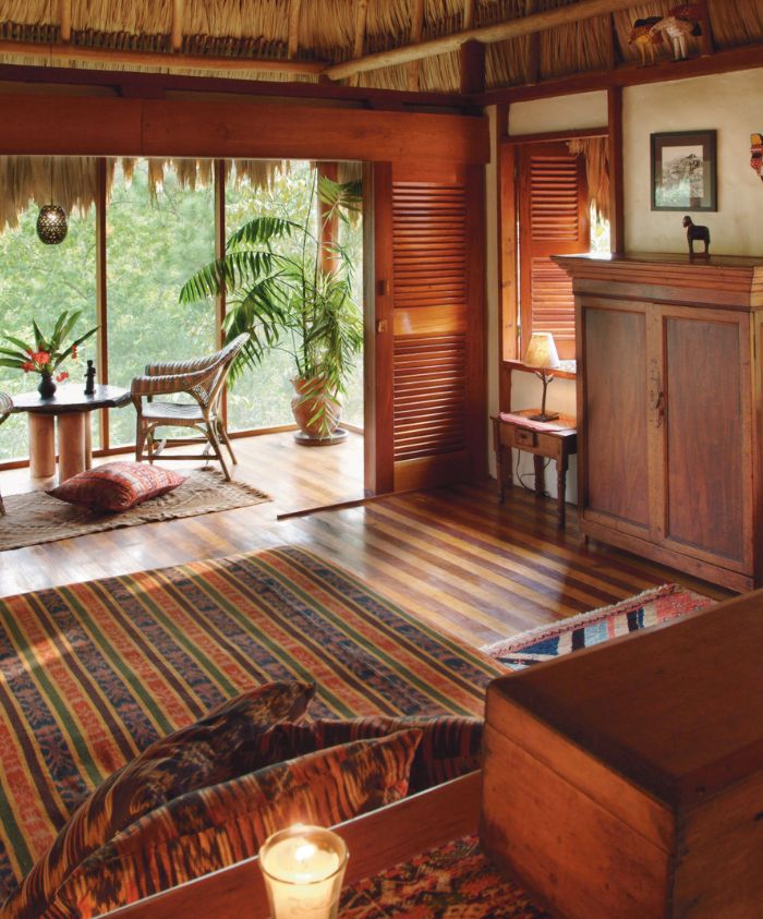 A cozy, rustic interior of a tropical cabin featuring wooden furniture, large windows with lush greenery outside, and colorful textiles on the floor. The space has a warm ambiance with natural light illuminating the room.