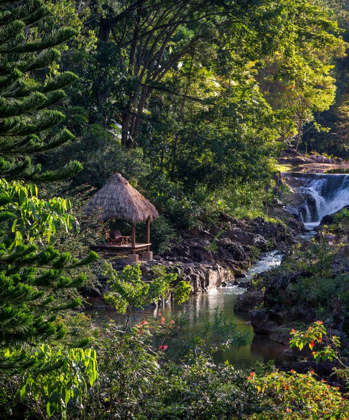 A serene landscape featuring a small thatched gazebo near a tranquil stream, surrounded by lush greenery and trees. In the background, a gentle waterfall cascades over rocks, creating a picturesque scene.