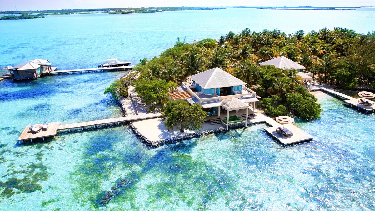 Aerial view of a tropical island resort featuring overwater bungalows, lush palm trees, and crystal-clear turquoise waters. Sandy pathways connect the bungalows, and sun loungers are visible on the deck.