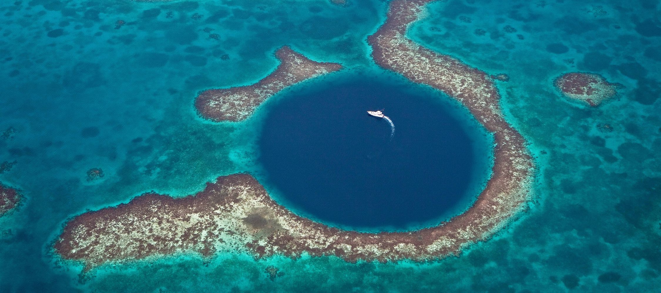 Aerial view of a circular blue hole in the ocean, surrounded by vibrant coral reefs and shallow turquoise waters. A small boat is navigating the water above the deep blue center.