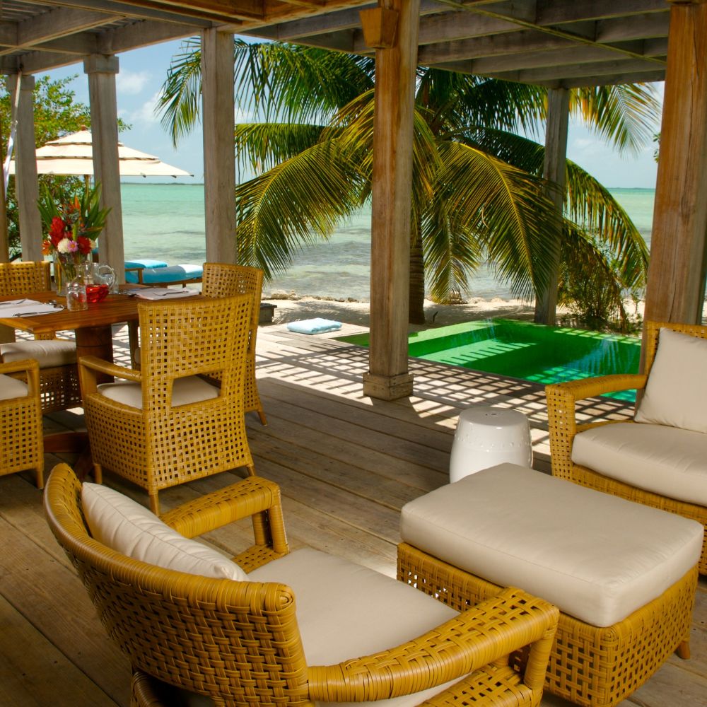 A tropical outdoor seating area with wicker furniture overlooks a serene beach. Palm trees frame the view, and a small pool is visible beside the seating. A dining table is set with flowers, creating a relaxing atmosphere.