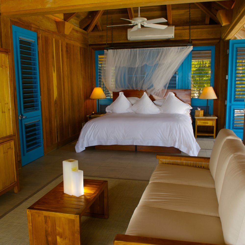 A cozy wooden bedroom featuring a king-sized bed with white bedding, two decorative pillows, and blue window shutters. There’s a comfortable beige sofa and a small wooden table with a decorative candle. The room has a tropical vibe with warm wood tones and a ceiling fan.