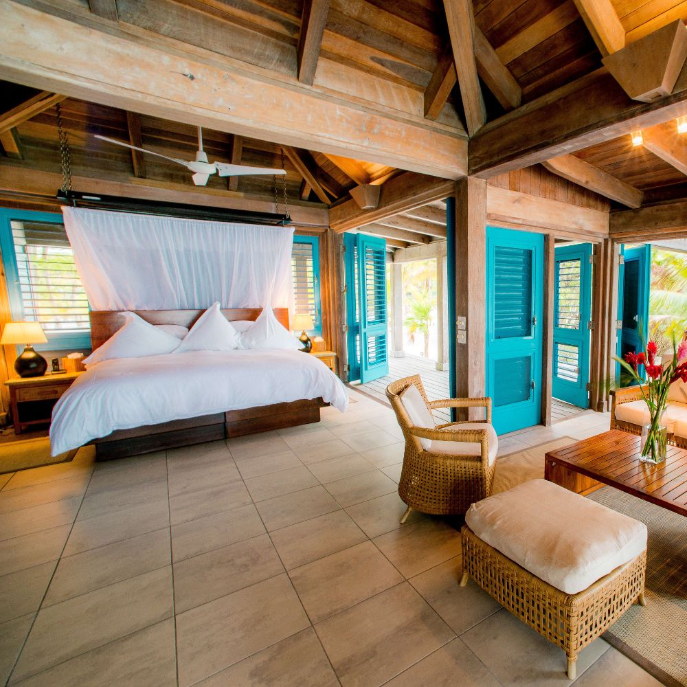 Bright and airy bedroom featuring a king-sized bed with white linens, a large rattan chair, and a coffee table. Blue shutters open to a tropical exterior, complemented by wooden beams and tiled flooring.