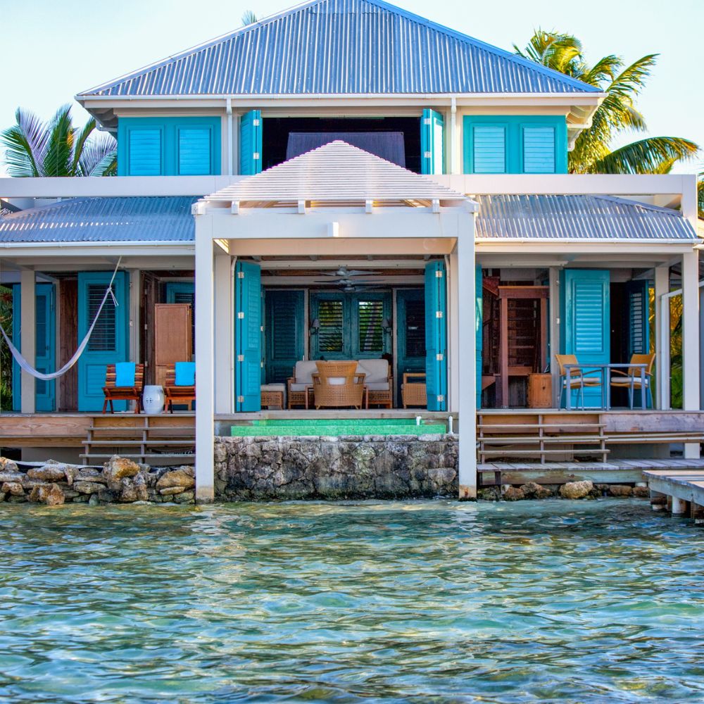 A coastal house with a wooden deck and blue shutters overlooks a clear, turquoise lagoon. There are lounge chairs and a hammock on the deck, surrounded by palm trees. The roof is metallic and sloped.