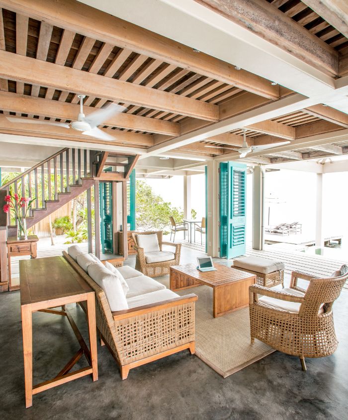 A stylish, open-plan living area featuring wooden beams and furniture. The space includes a comfortable white sofa, rattan chairs, and a wooden dining table. Large windows with teal shutters provide a view of the outdoor area.
